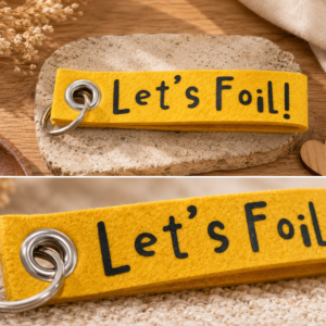 Gelber Filz-Schlüsselanhänger mit Let’s Foil Schriftzug auf dekorativem Untergrund