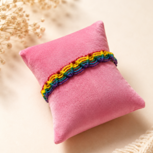 Handgeknüpftes Regenbogen-Makramee-Armband aus buntem Baumwollgarn auf rosafarbenem Schmuckkissen.