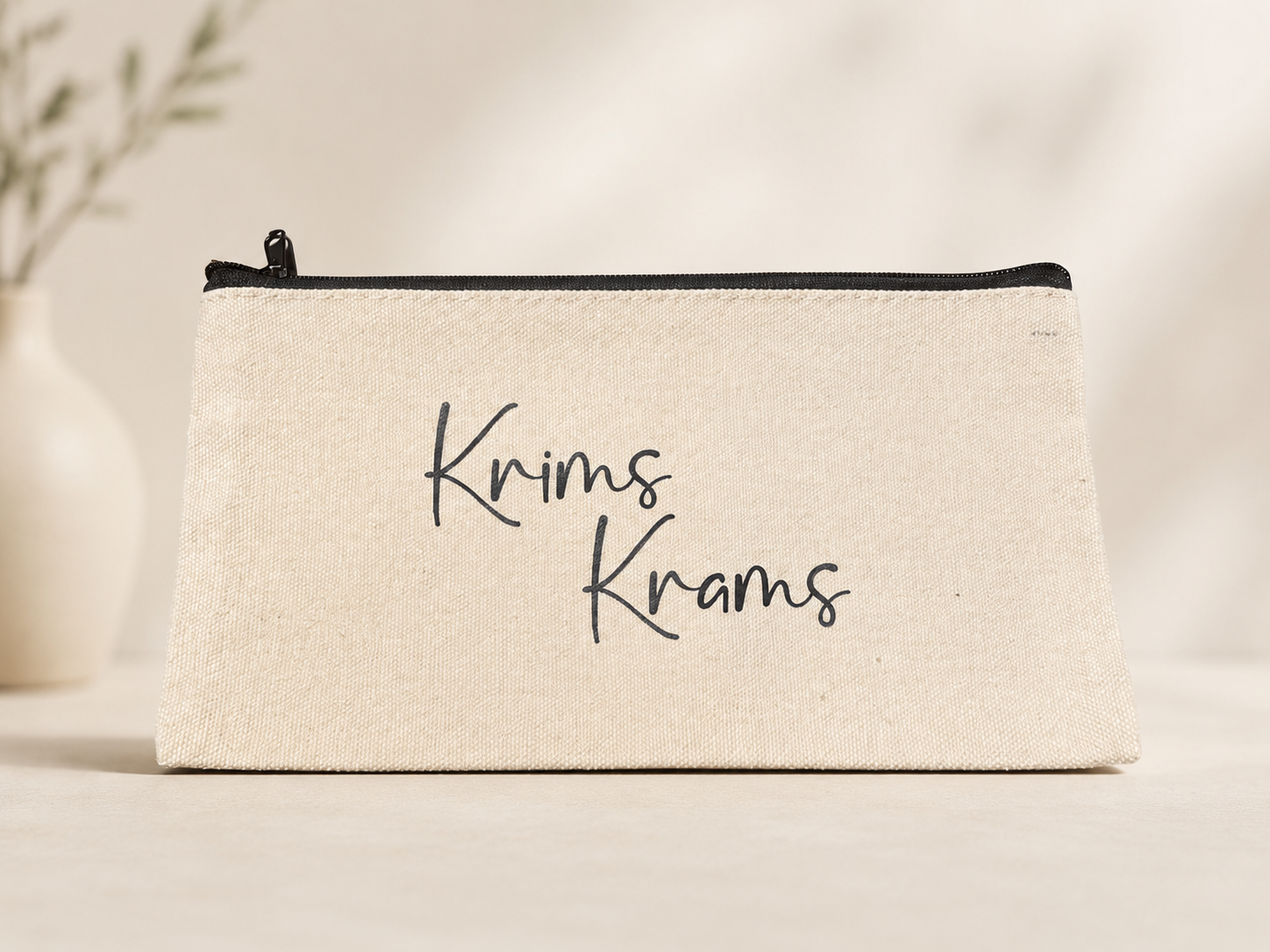 Naturfarbene Kosmetiktasche mit schwarzem Reißverschluss und Aufdruck „Krims Krams“ auf hellem Hintergrund.